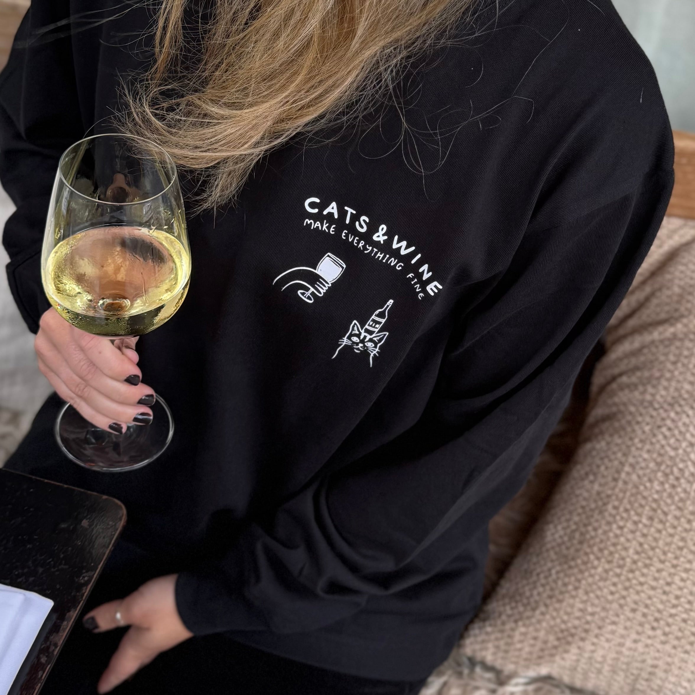 Person wearing a black sweatshirt with 'Cats & Wine' text and graphics, holding a glass of white wine.