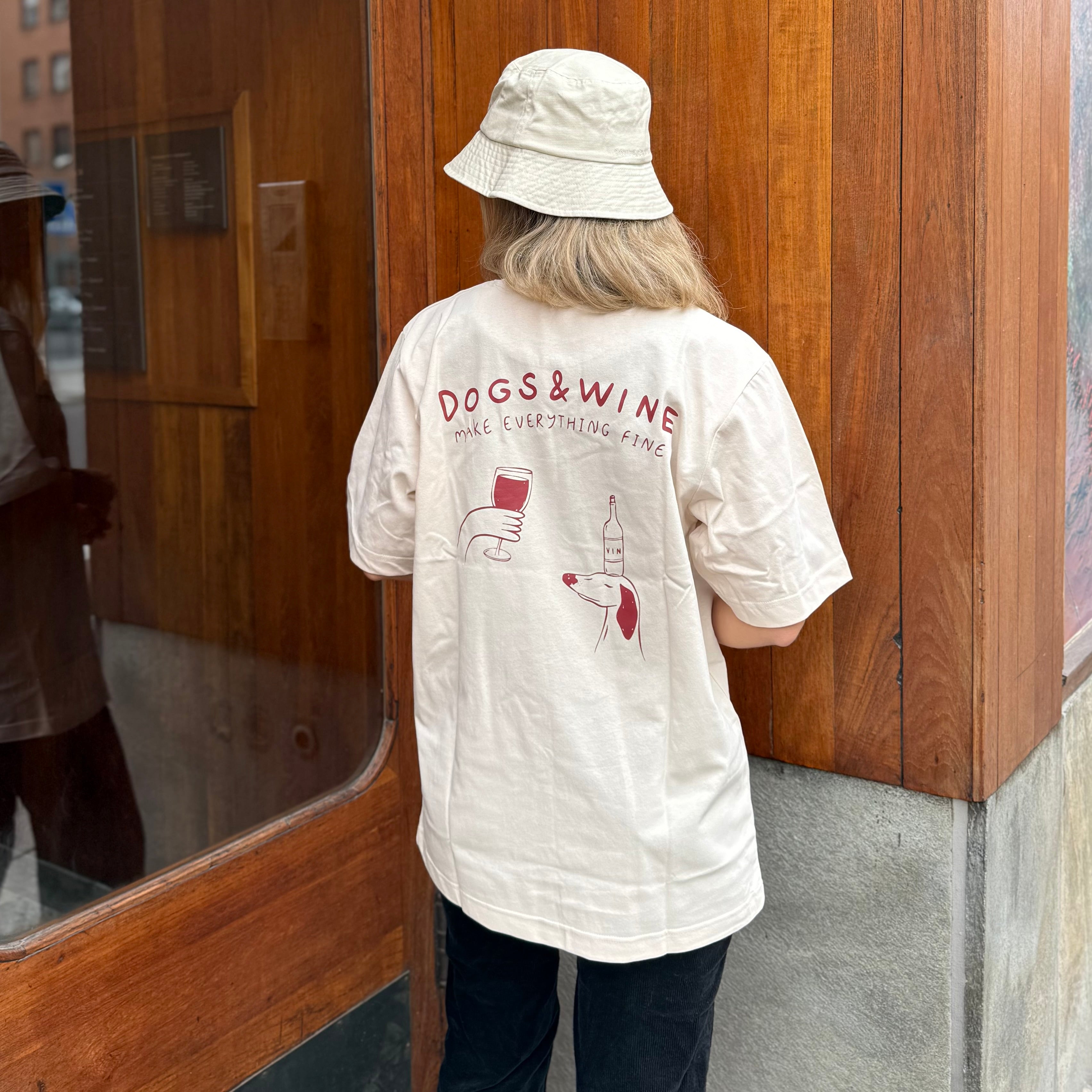 Person wearing a white shirt with 'DOGS & WINE' text and graphics, standing in front of a wooden door.